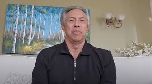 A man with gray hair sits on a white couch in front of a wall with a painting of birch trees and a light fixture.