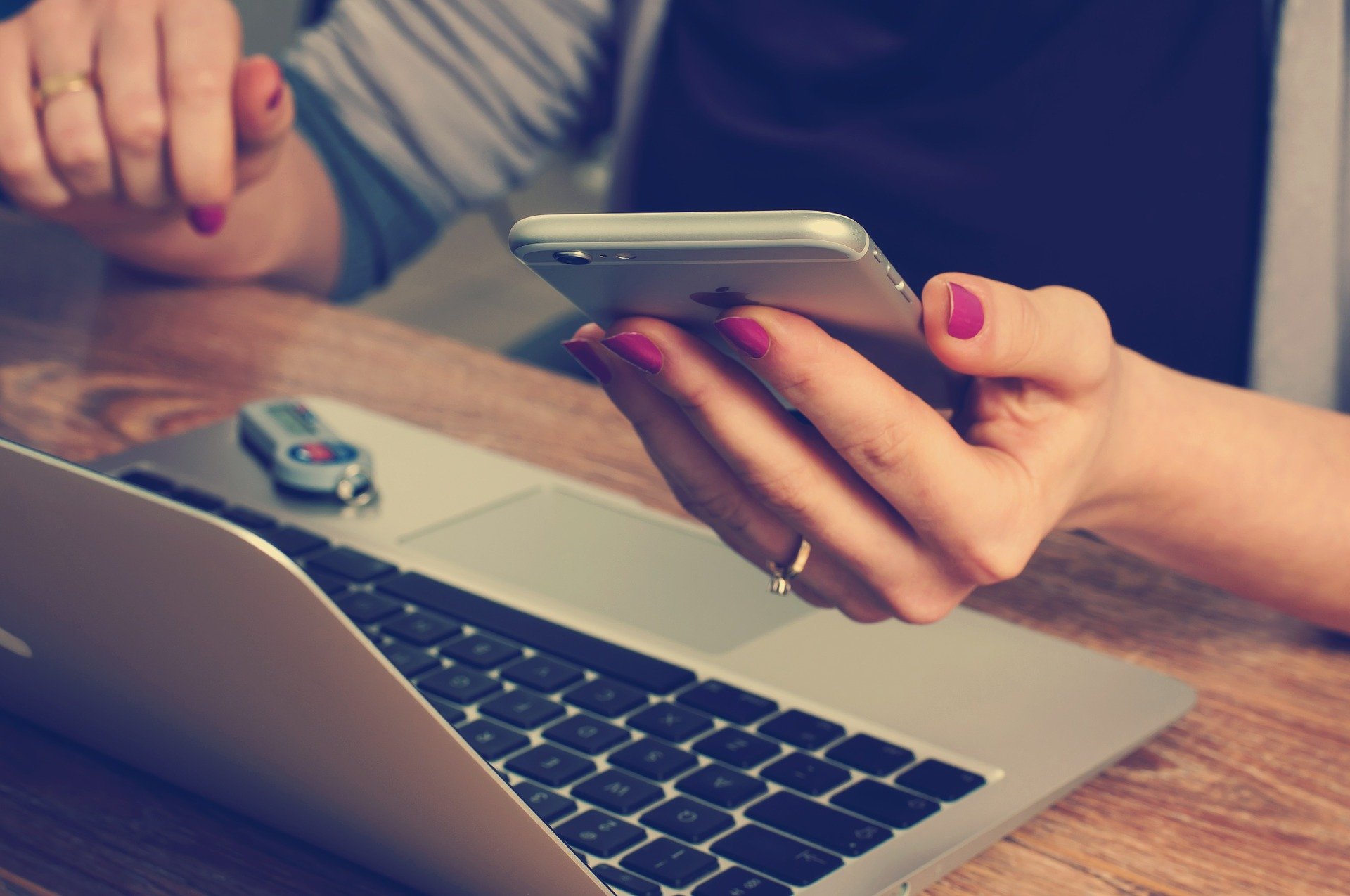A woman looking at an Iphone with a laptop on top of her table