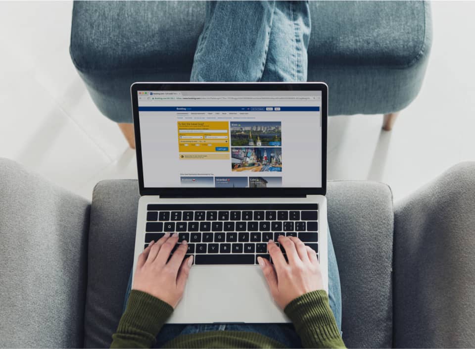 Person sitting on a chair with legs up on an ottoman, using a laptop displaying a travel booking website.
