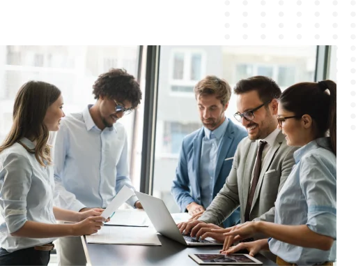Five people in business attire stand around a table in an office, collaborating with documents, a laptop, and a tablet.