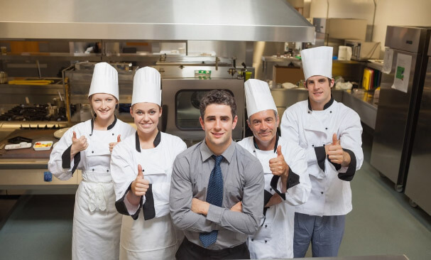 Four people in chef uniform doing thumbs up and one in long sleeves and tie inside a kitchen
