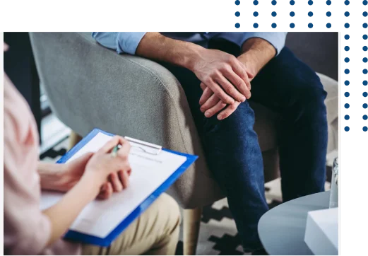 Two people sit facing each other; one holds a clipboard and pen, appearing to take notes, while the other sits with hands clasped, suggesting a counseling or therapy session.