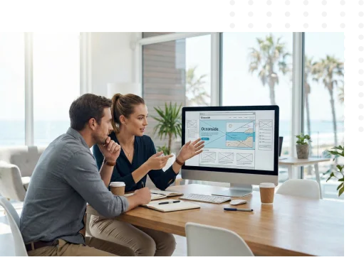 Two people sit at a desk with coffee, discussing a website design displayed on a large computer screen in a bright office with palm trees visible outside the window.